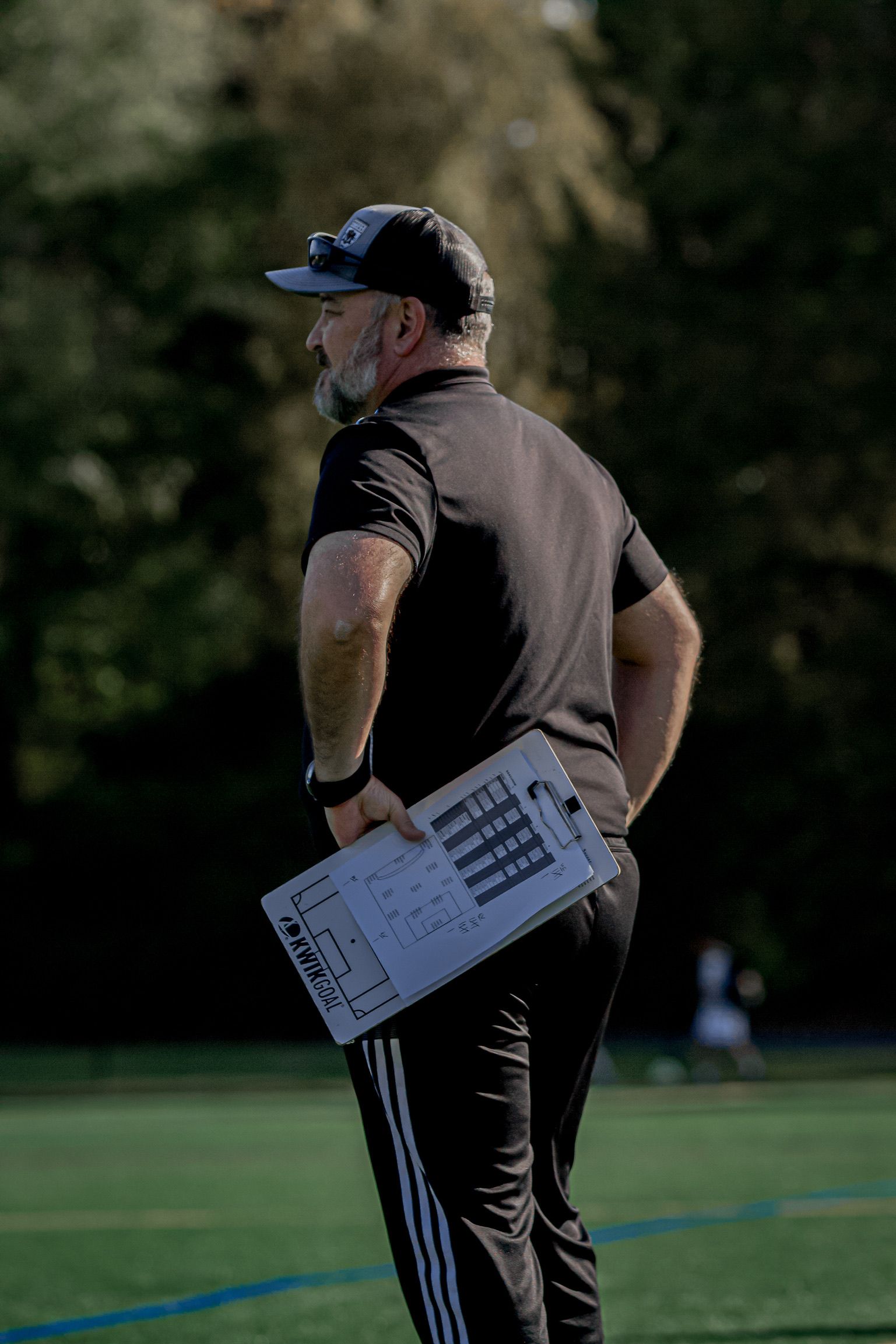 Coach in black sportswear holding a clipboard on a football field, seen from behind.