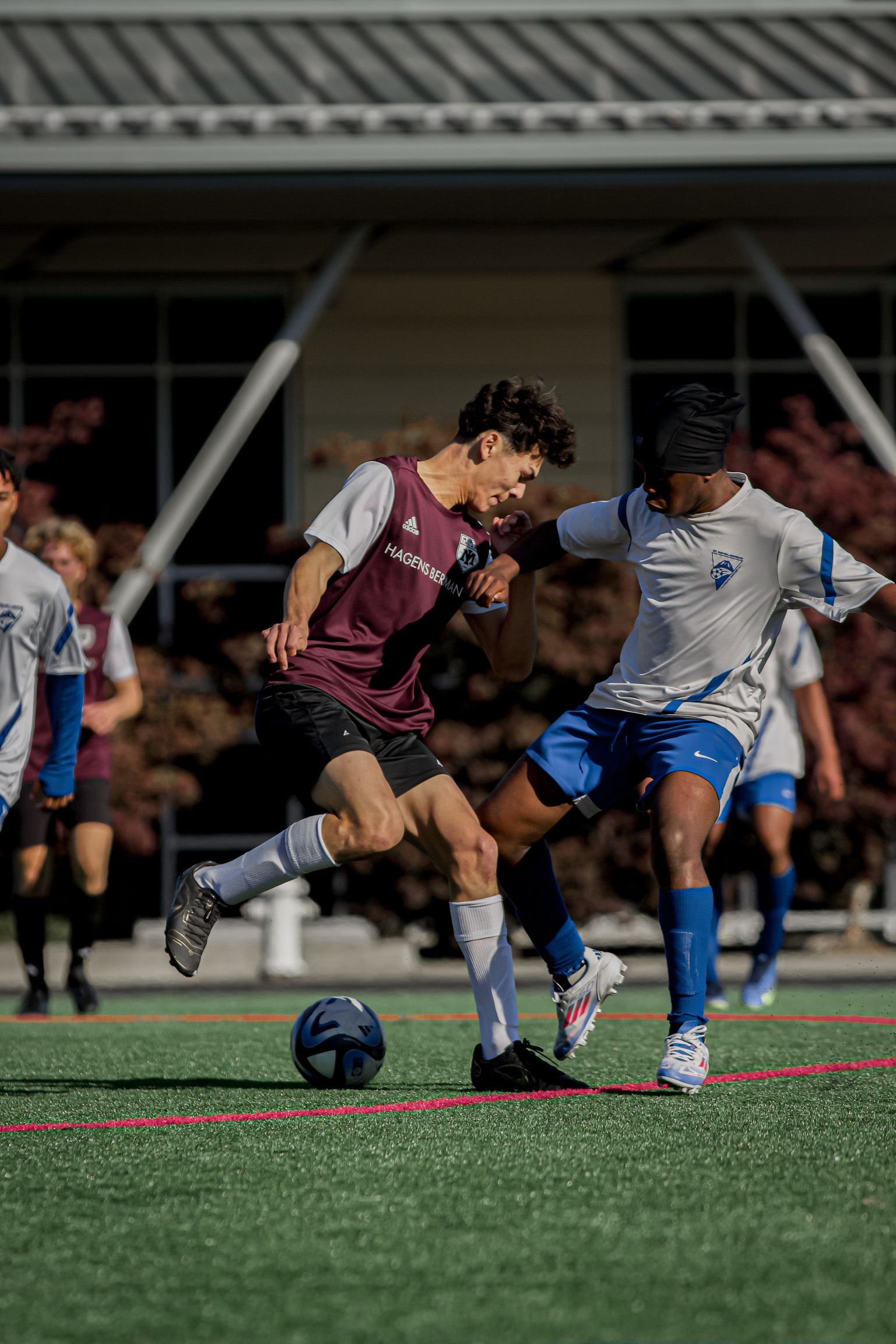 Two soccer players battle for the ball on a field, one in maroon and one in blue and white.