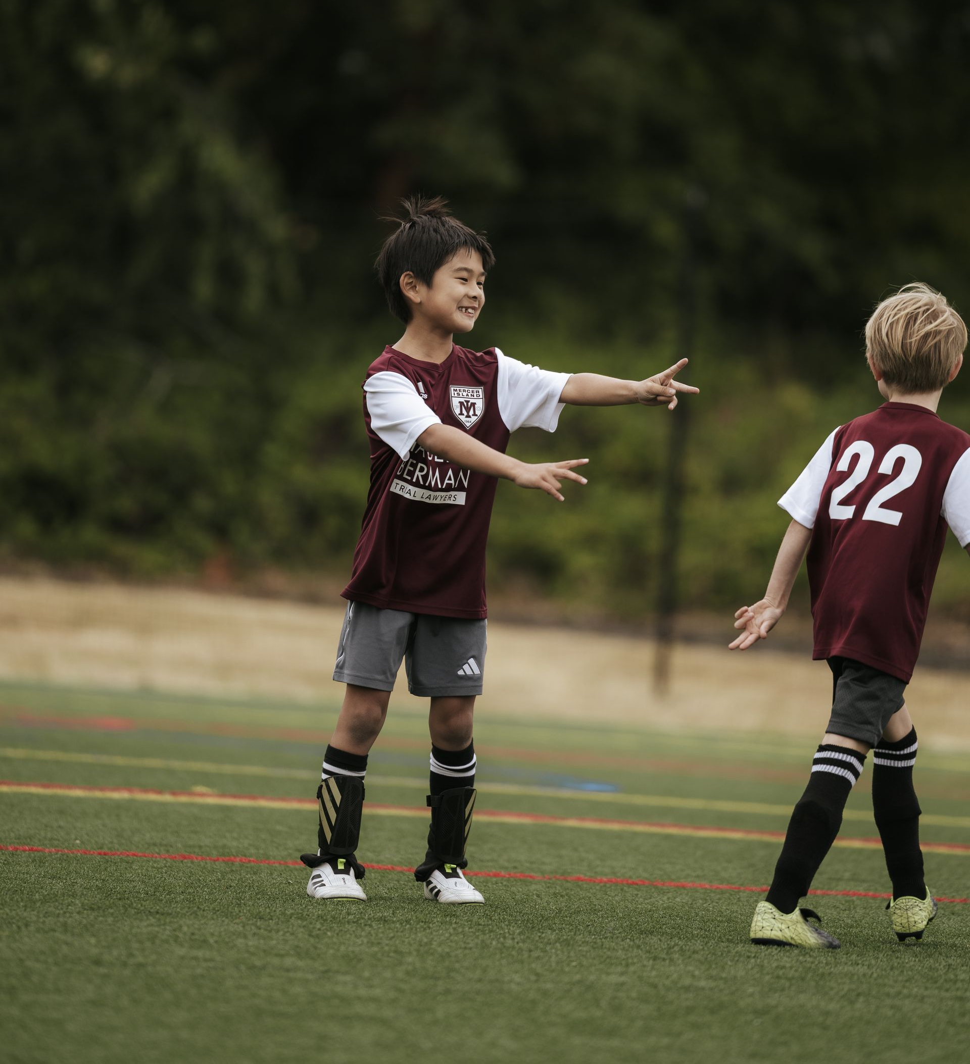 Two young soccer players in maroon jerseys on a field, one reaching out to the other during a game.