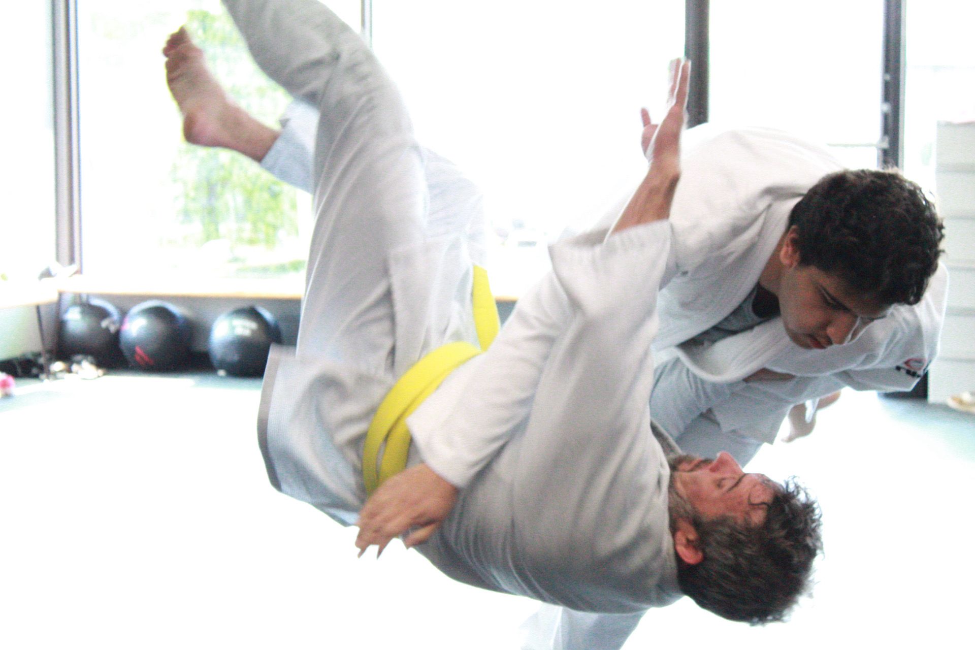 Two people practicing judo, one throwing the other in a gymnasium on a yellow mat.