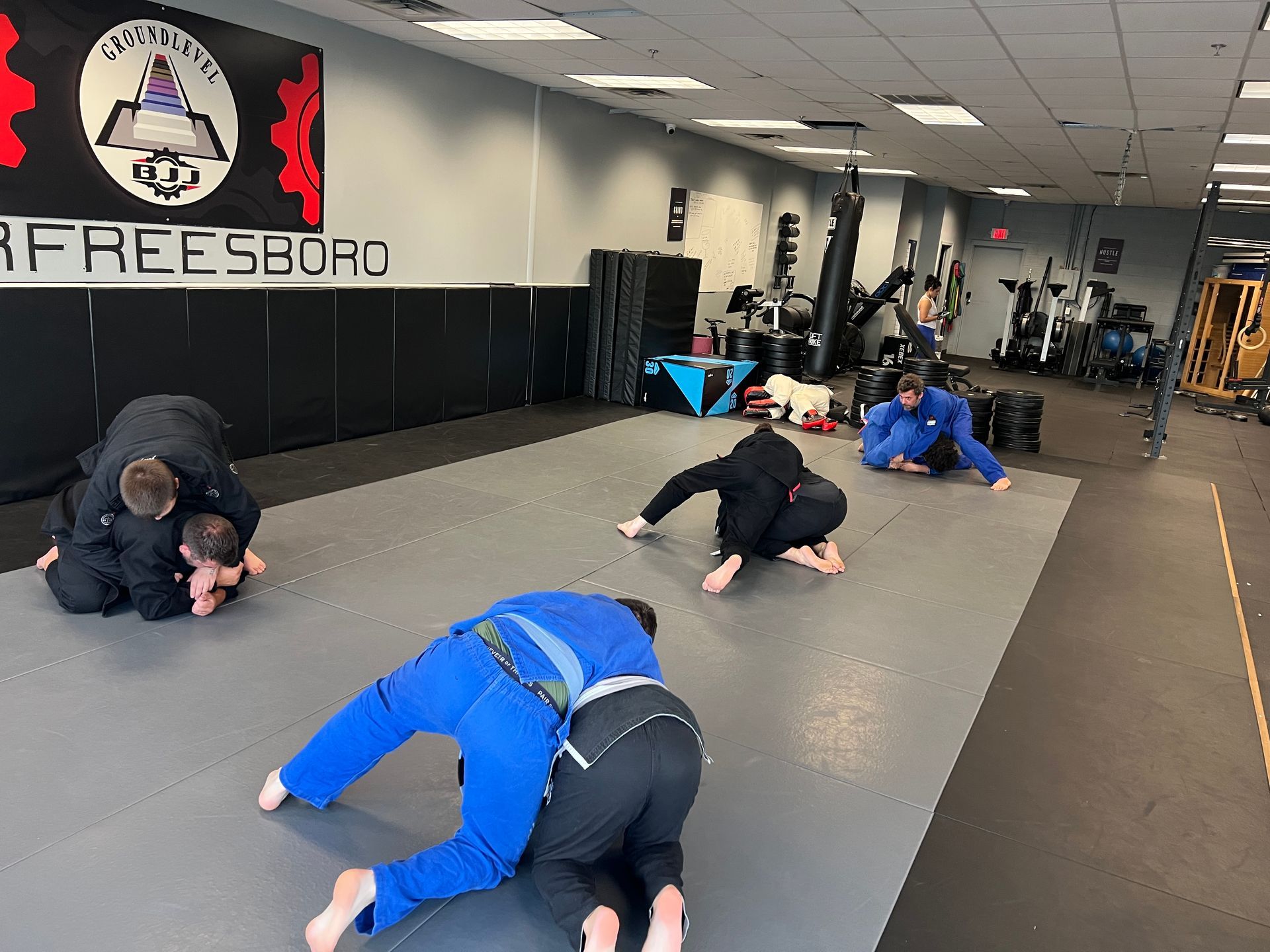 Two wrestlers grappling on a gym mat while a trainer watches in the background.