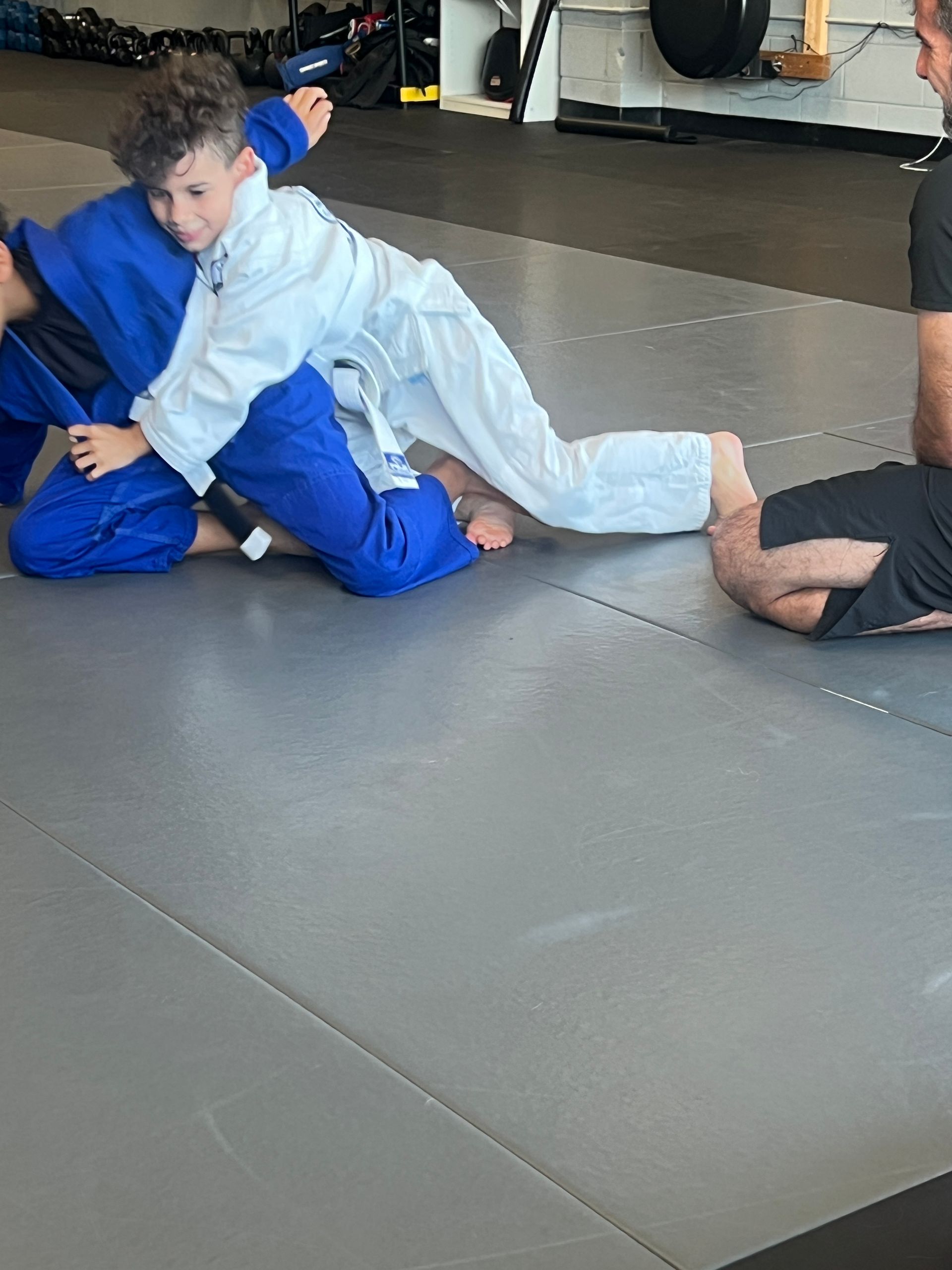 Two martial arts students in white and blue gis face off, with three seated observers in the background.