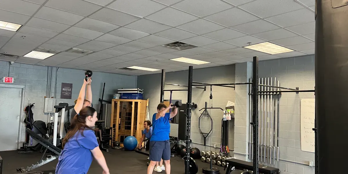 Two people in blue shirts do overhead exercises in a gym with weights and equipment.
