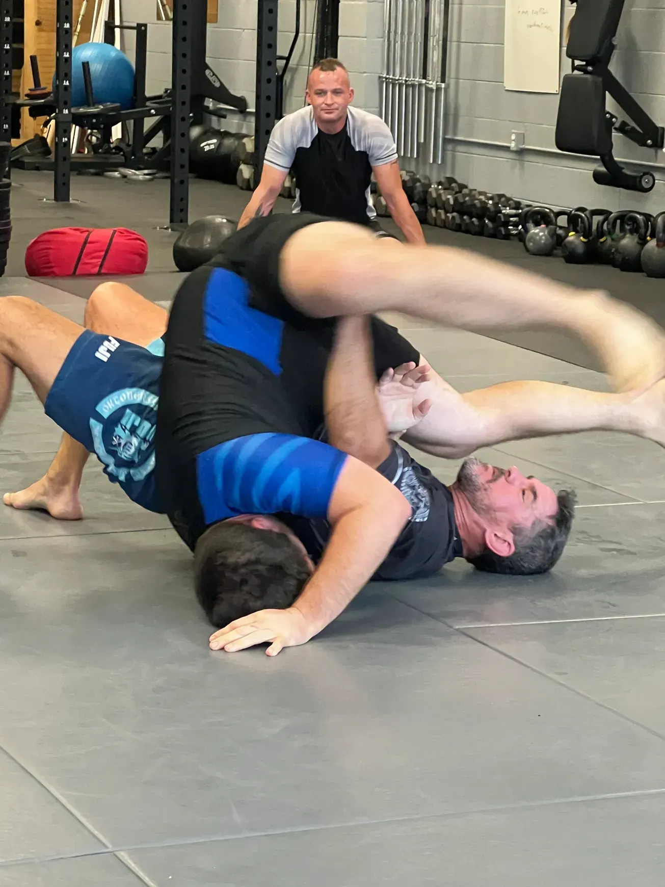 Two wrestlers grappling on a gym mat in Murfreesboro, TN while a trainer watches in the background.