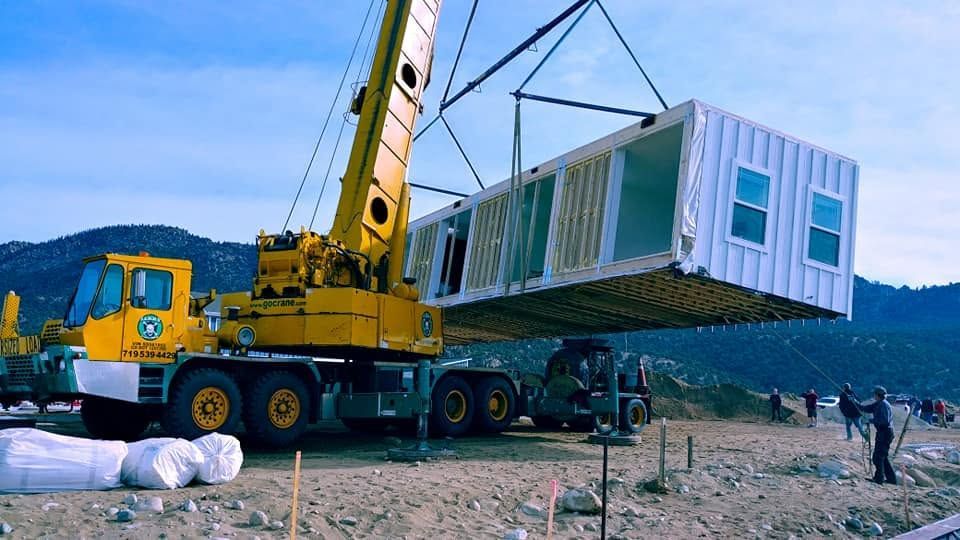 Yellow crane lifting a white modular house onto a site in a rocky landscape under a blue sky