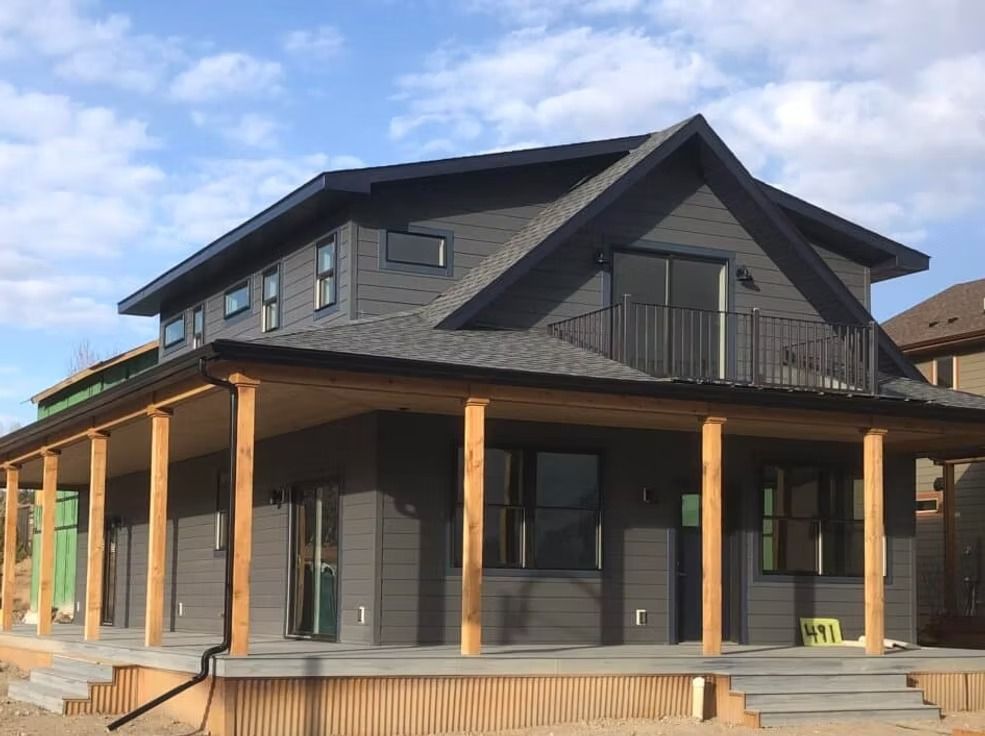 Modern two-story gray house with a wraparound porch and wooden columns under a blue sky