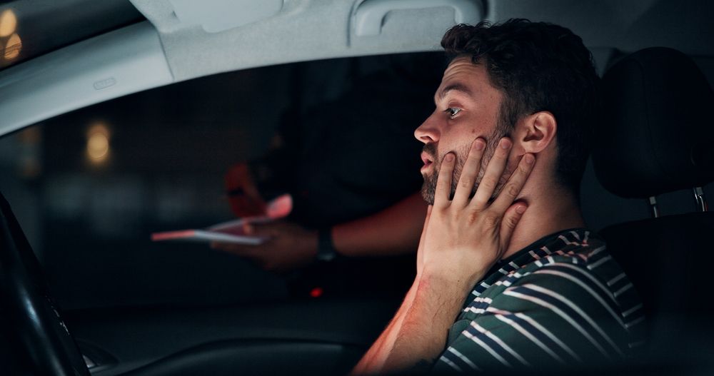 A person in a striped shirt sits in a car, hands on their face, looking startled as an officer holds a document outside.