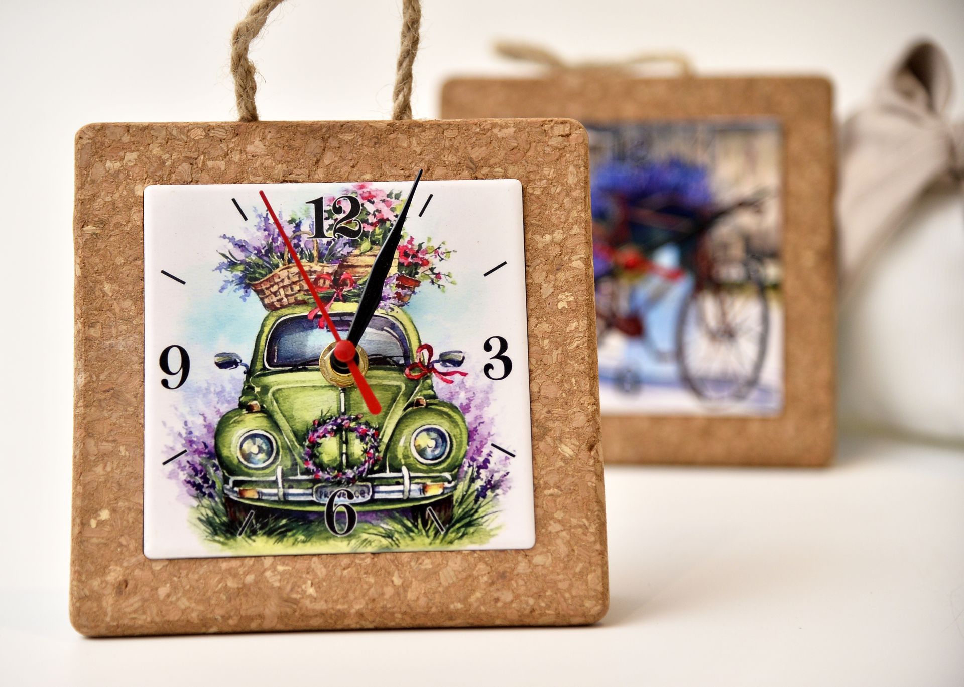Un orologio quadrato con cornice in sughero raffigura un'auto verde con dei fiori sul quadrante; dietro c'è una seconda foto.
