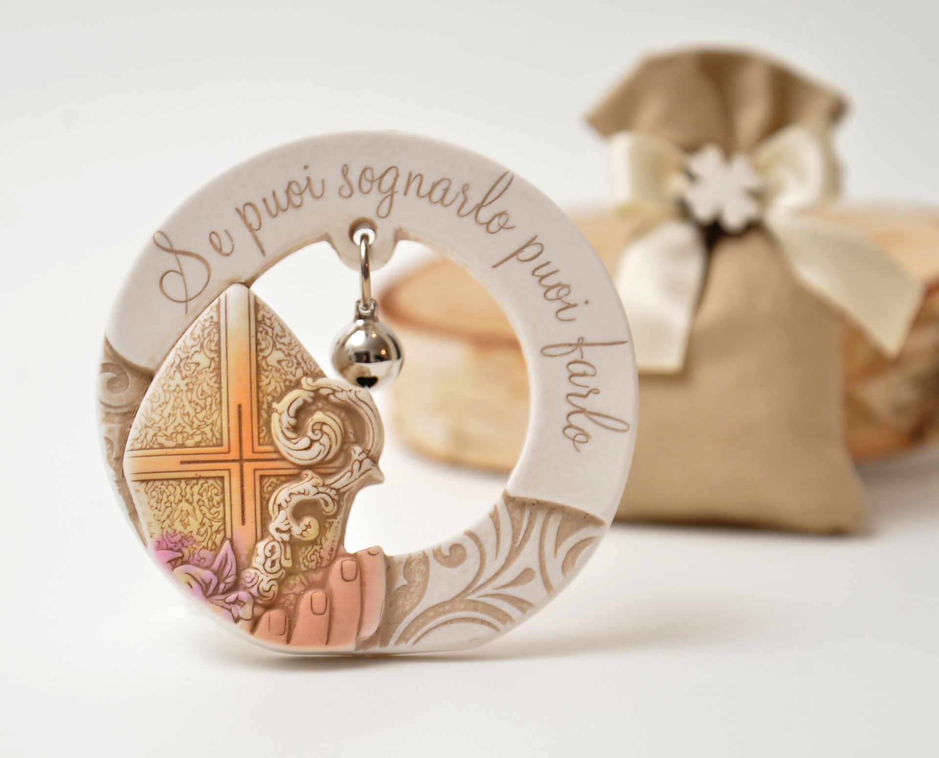 Ciondolo in ceramica con l'immagine di un vescovo e testo in italiano, una piccola campanella e una borsa in tessuto beige sullo sfondo.