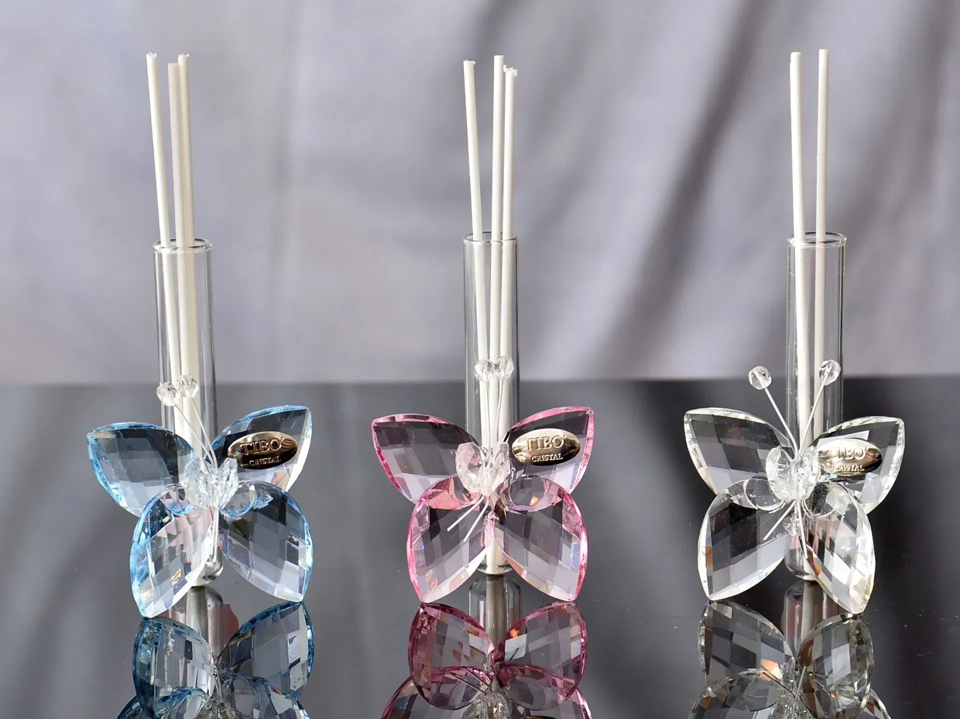 Tre diffusori di aromaterapia a forma di farfalla di cristallo, nei colori blu, rosa e trasparente, con tubi di vetro e bastoncini.