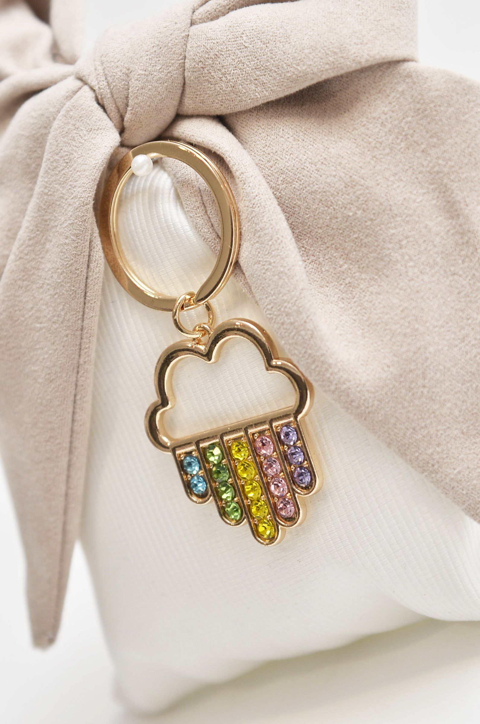Ciondolo a forma di nuvola con pioggia arcobaleno attaccato a un fiocco beige annodato attorno a una pochette bianca.