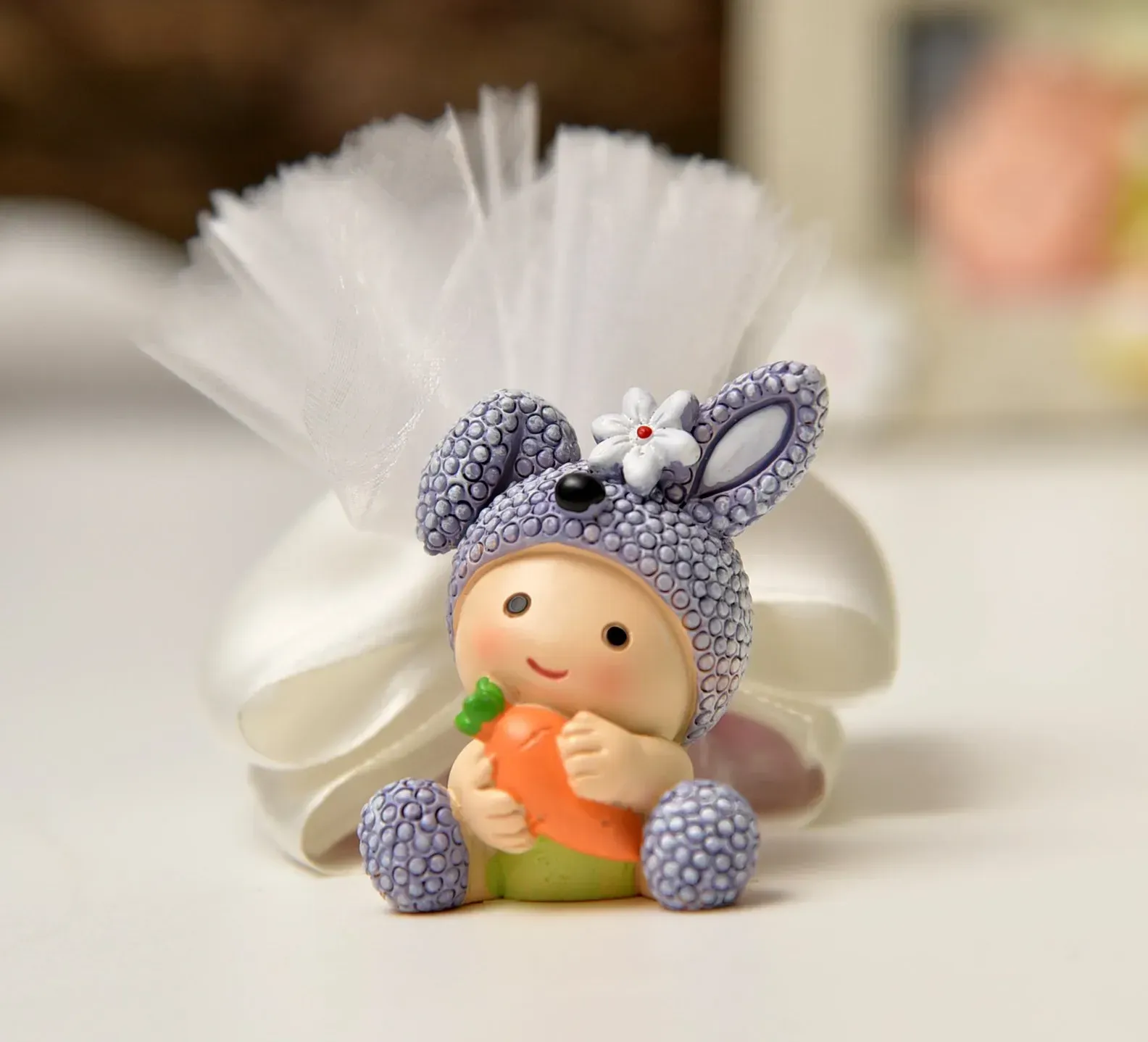 Una piccola statuetta di un bambino con un cappello da coniglio, che tiene in mano una carota, davanti a una borsa di tulle bianco.