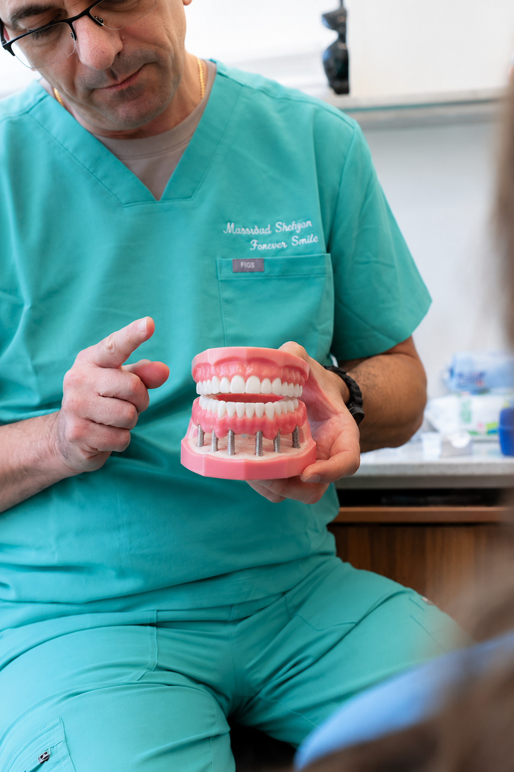 Dentist in teal scrubs holding pink dental models and pointing at the teeth in a clinic.