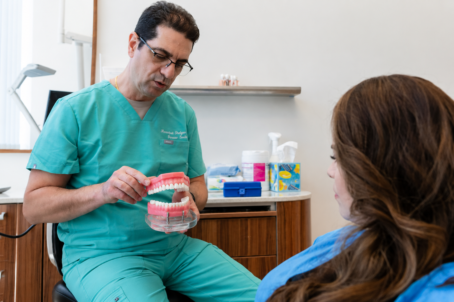Dentist in green scrubs shows a dental model to a seated patient in a clinic office.