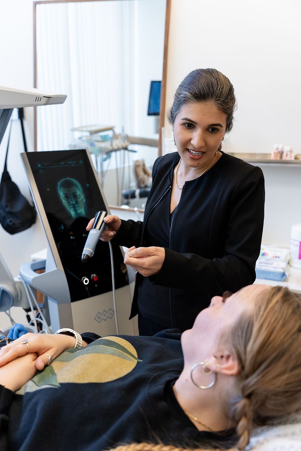 Clinician pointing at a medical display in a dental office, with equipment and a seated patient in view