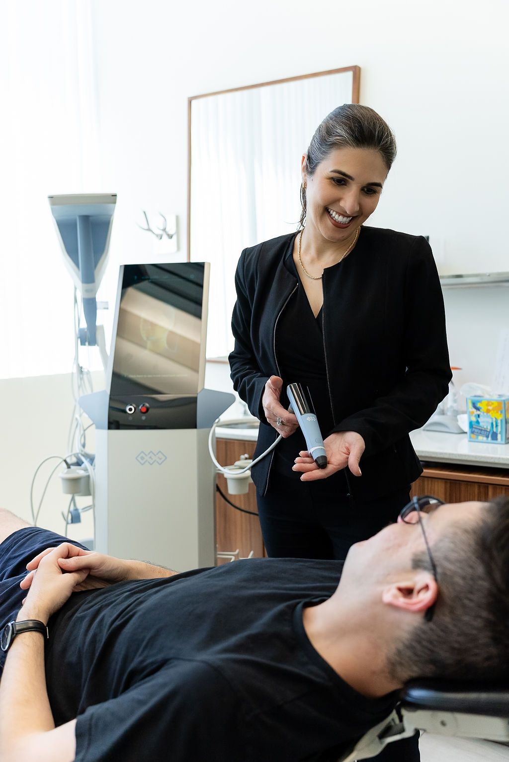 Woman in black uniform performing a cosmetic treatment on a reclining client in a clinic room
