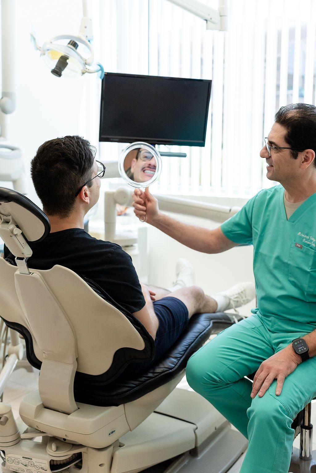 Dentist in green scrubs showing a patient a mirror during a dental exam in a bright clinic
