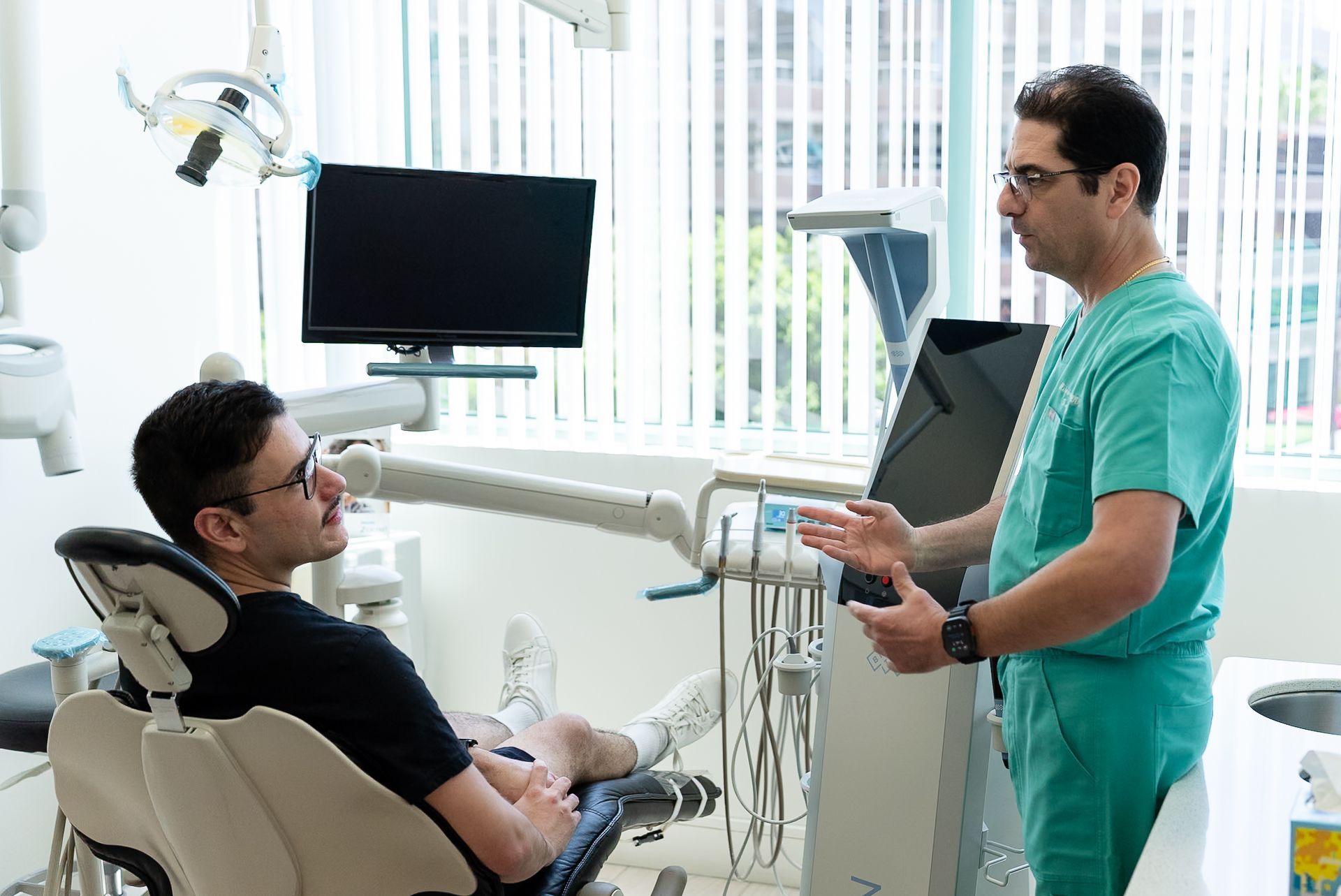 Dentist in green scrubs speaking with seated patient in a bright dental clinic