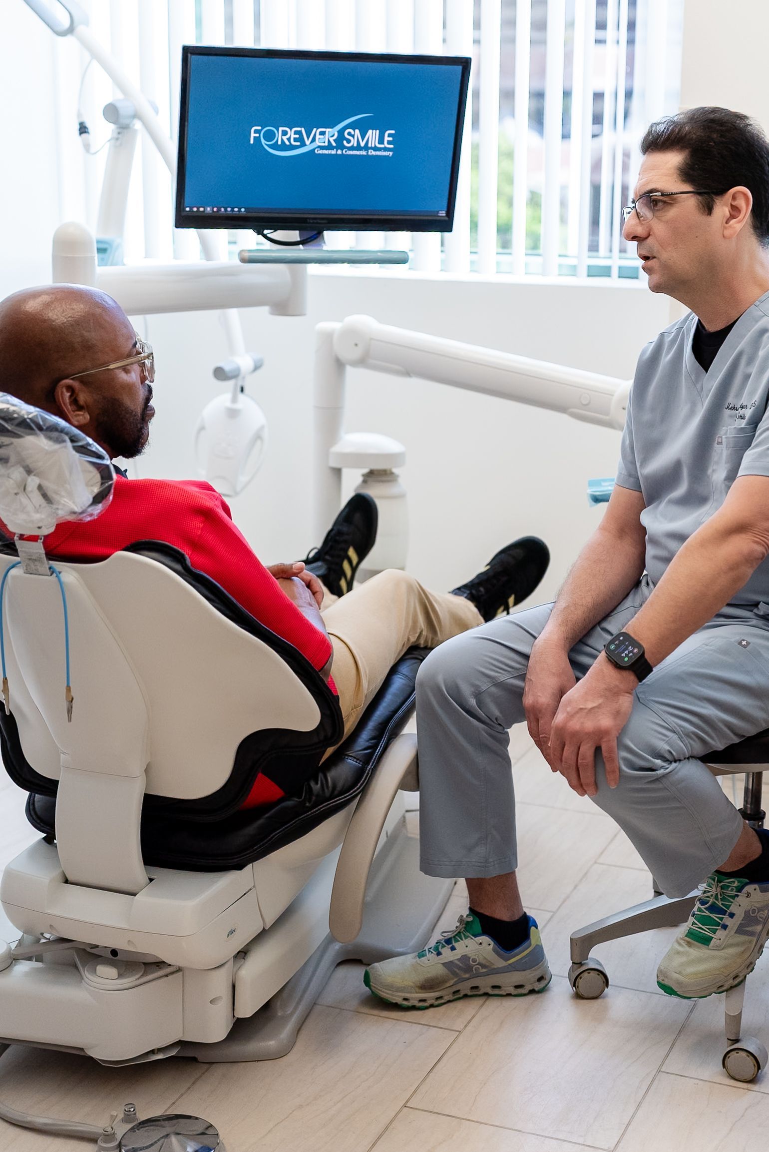 Patient in a red jacket speaks with a clinician beside a medical exam chair and monitor in a clinic