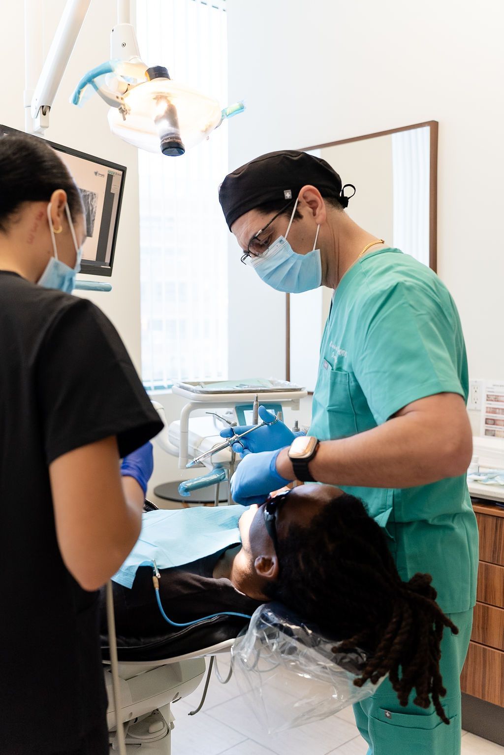Dentist in green scrubs treating a patient in a dental clinic, with an assistant nearby.