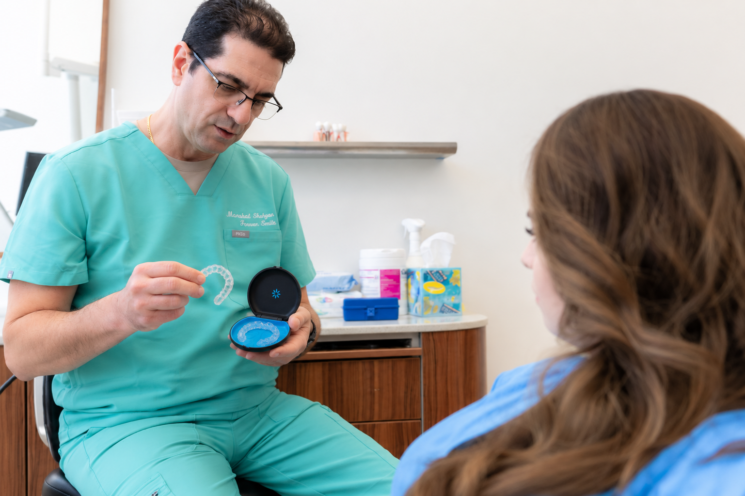 Dentist in green scrubs showing a dental model to a seated patient in a clinic room