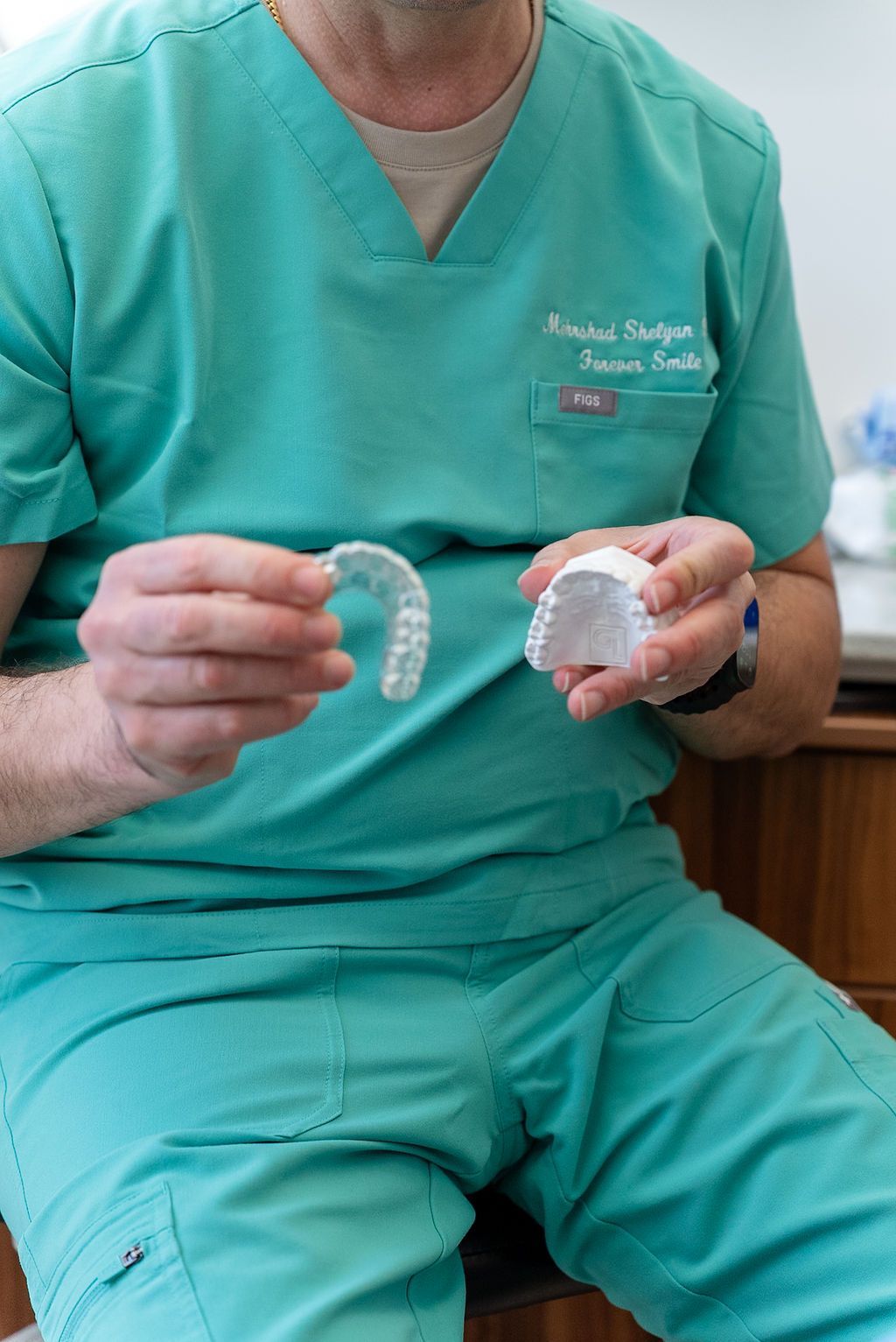 Dental professional in green scrubs holding two clear dental aligners while seated indoors