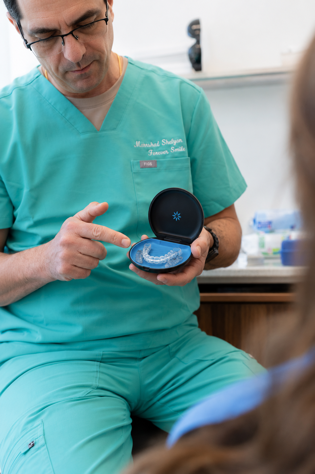 Doctor in green scrubs showing a small open case with a blue dental appliance to a seated patient