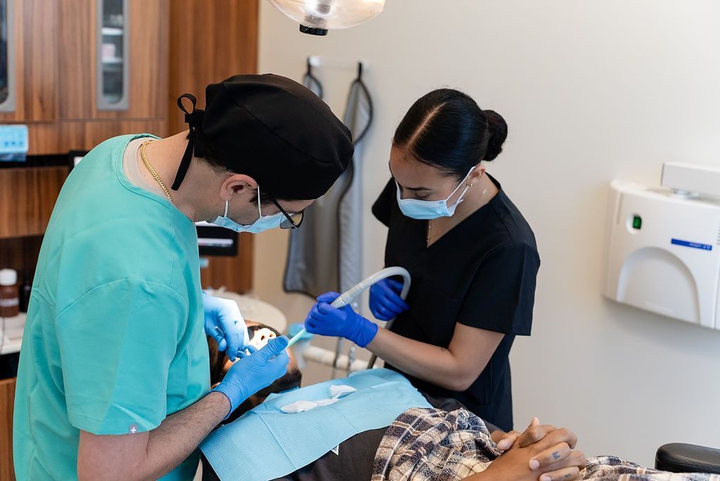 Dentist and assistant treating a reclining patient in a dental clinic, both wearing masks and gloves.