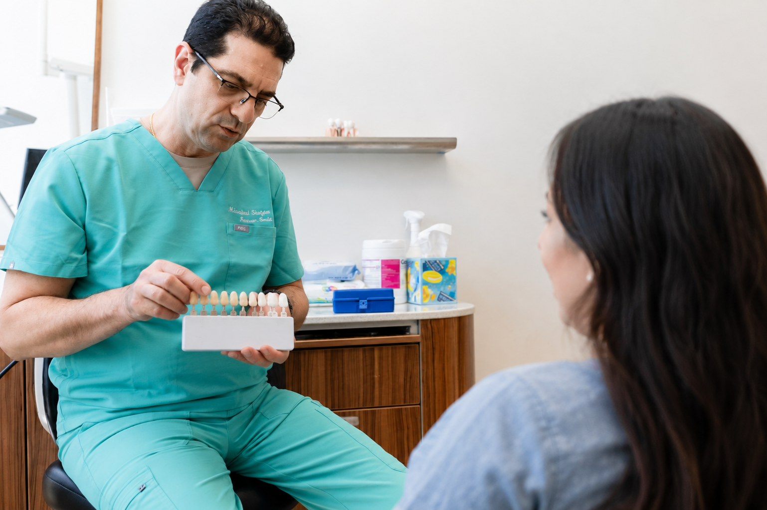 Dentist in green scrubs speaking with a seated patient in a bright dental clinic
