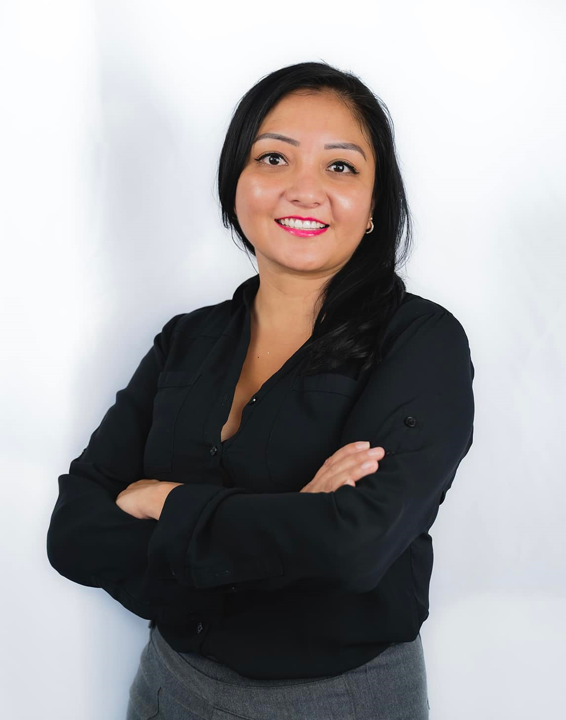 A person with dark hair, wearing a black long-sleeved shirt and gray skirt, stands smiling with arms crossed.