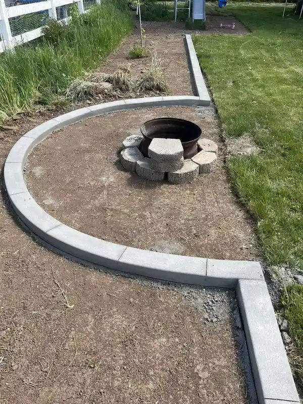 Curved concrete garden border around a small stone fire pit on a dirt patch beside green lawn.