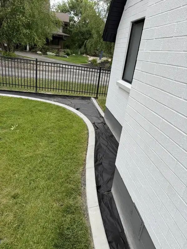 Curved lawn beside white house with black drainage liner along foundation and metal fence in background