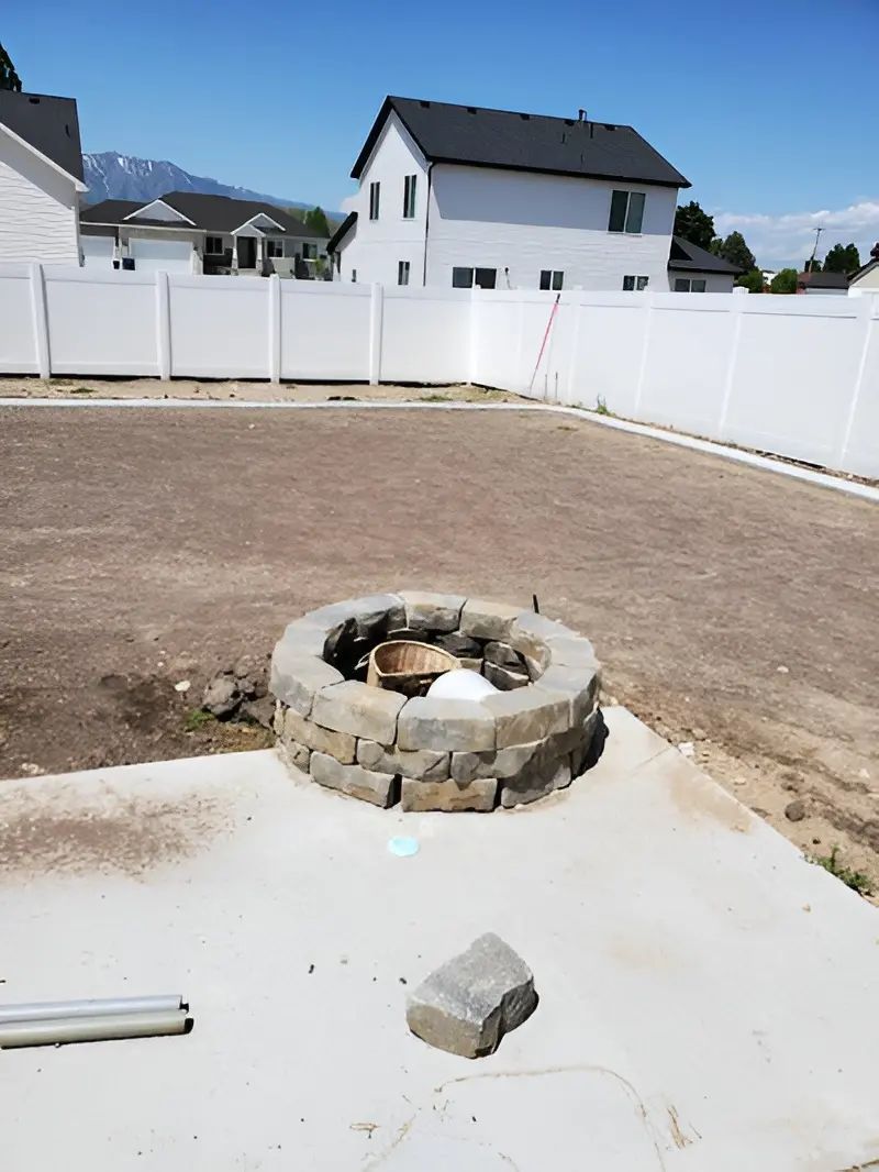 Backyard with a stone fire pit, concrete patio, and white fence behind houses.