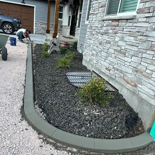 Curved garden bed with dark rocks, small shrubs, and drainage grate beside a stone house wall