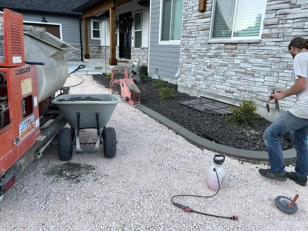 Worker spreading gravel beside a house, with a wheelbarrow and orange equipment on a stone walkway.