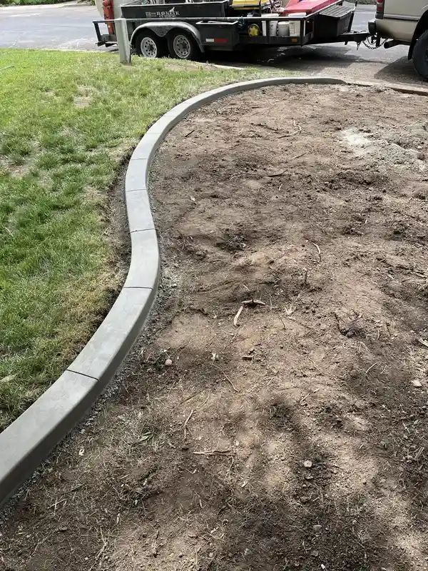 Curved concrete curb beside a dirt patch and grassy edge, with a truck trailer in the background