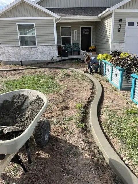 Concrete garden edging in a yard beside a house, with a wheelbarrow of soil in the foreground.