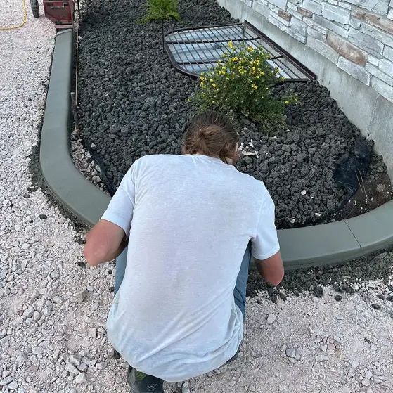Person kneeling on gravel beside a curved stone border in a landscaped yard, tending plants near a house