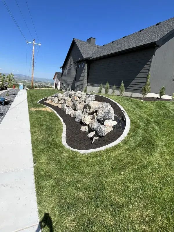 Curved rock garden with black edging beside a sidewalk and dark house on a sunny residential street