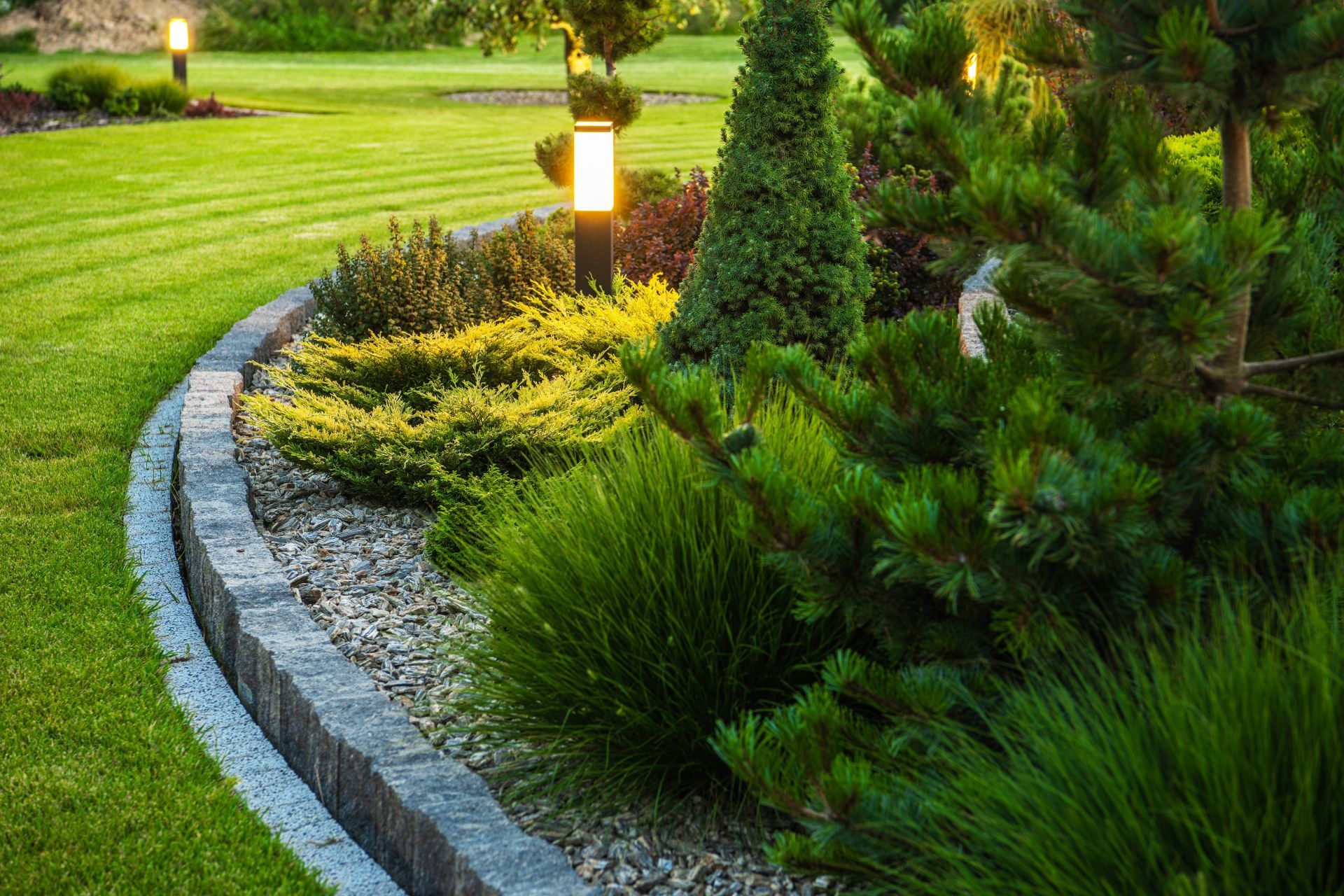 Curved garden path with stone edging, shrubs, and glowing lamp posts at dusk