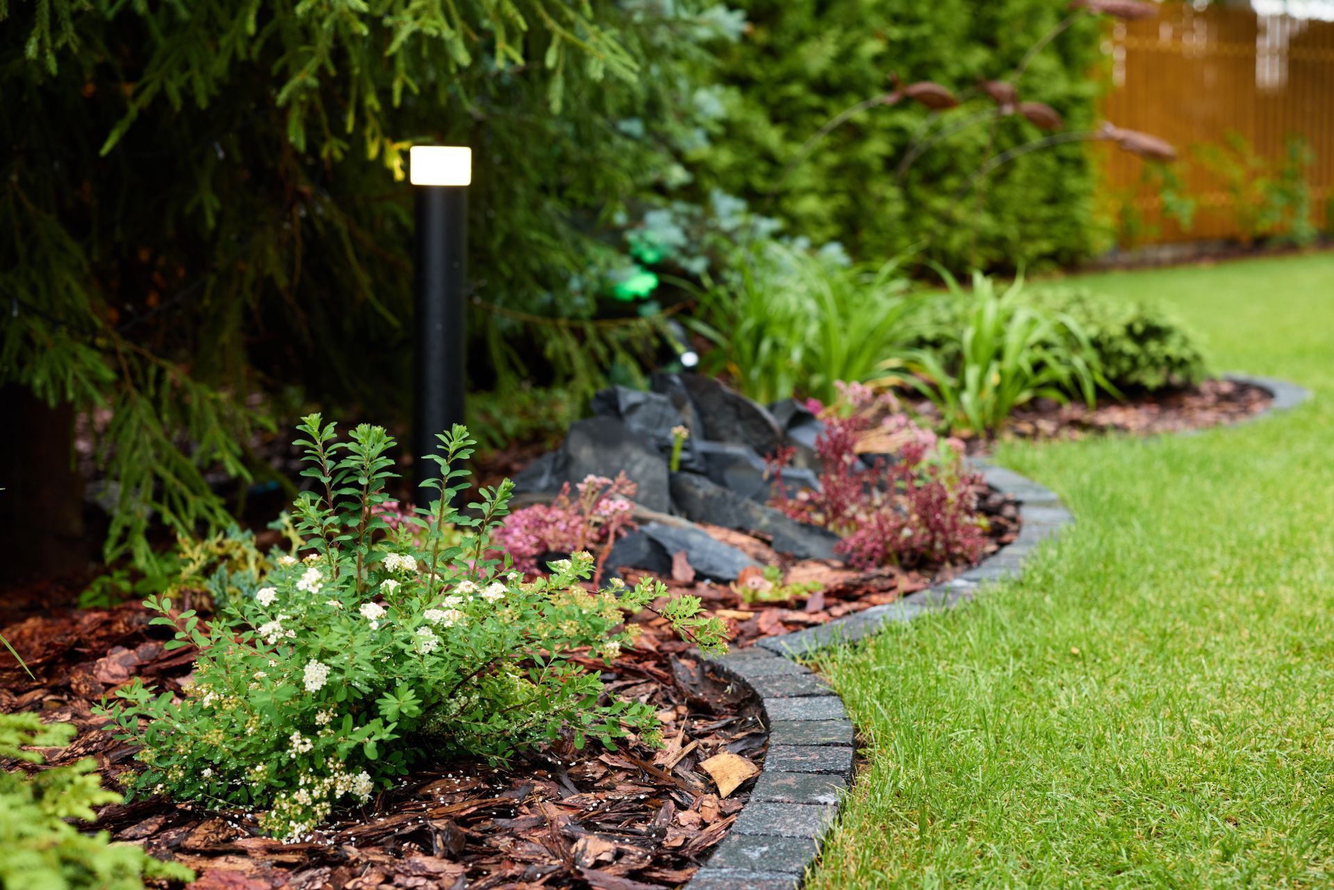 Curved garden border with flowers, rocks, and a lit black path light beside a green lawn