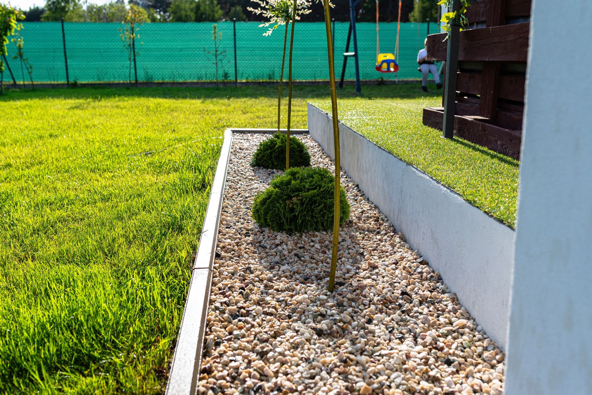 Landscaped gravel bed with small shrubs beside a white wall and green lawn under a fenced yard