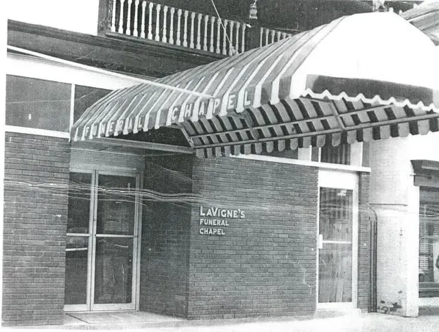 Black-and-white photo of a brick building entrance with striped awning and sign reading