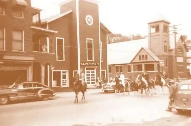 Sepia street scene with a brick building, parked cars, and people gathered near a church entrance