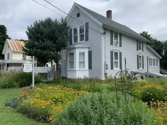 Gray house with white trim and a flower garden beside a sidewalk and signpost
