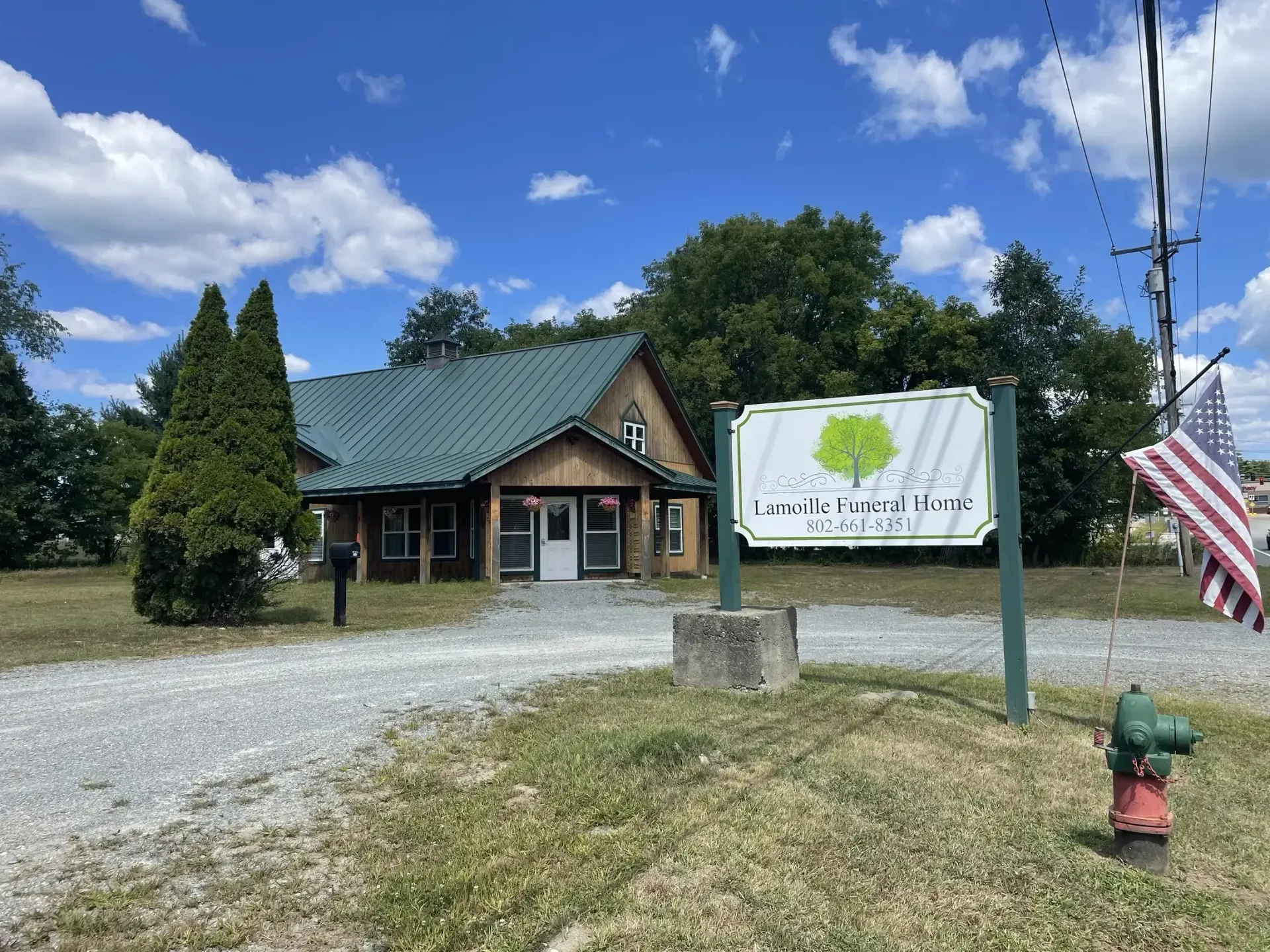 Small rustic building with green roof and sign, gravel lot, fire hydrant, and American flag under blue sky.