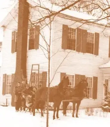 Horse-drawn sleigh in front of a snow-covered house with brown shutters, winter scene