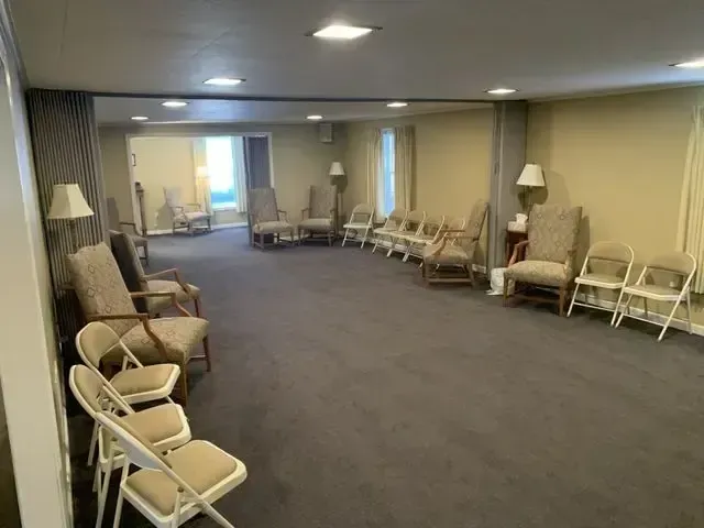 Empty hallway with rows of beige chairs and floor lamps along the walls, carpeted floor, and ceiling lights