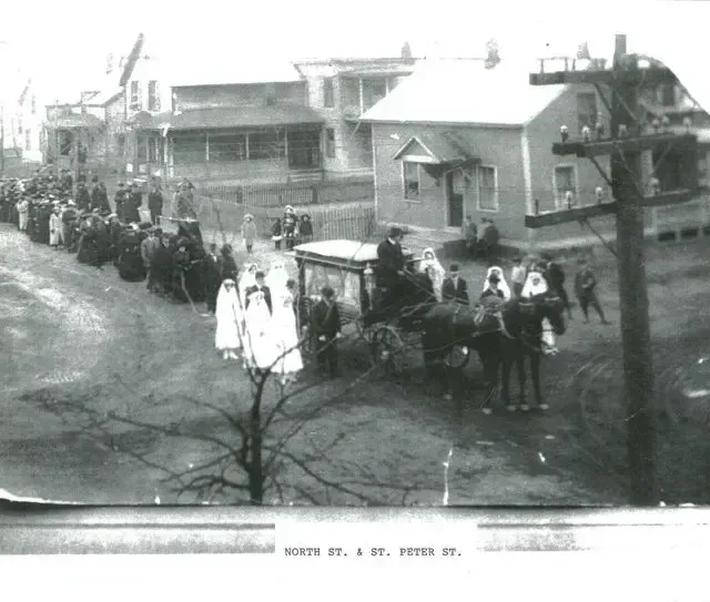 Crowd gathered on a street beside damaged buildings, with people standing and walking near a cart and house.