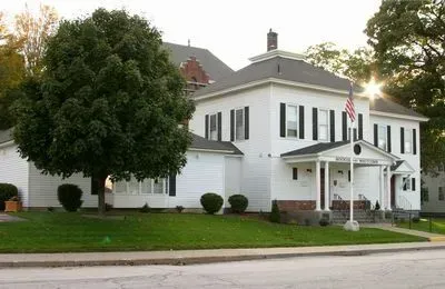 White two-story house with black shutters and a front porch beside a large tree on a suburban street
